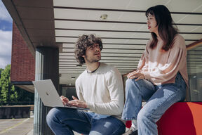 Two students outside a brick building, one using a silver laptop while talking with the other on a red bench