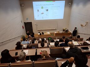 Lecture hall with students watching a man present slides on electrically heated textiles at the front
