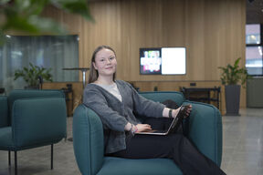 A person sits in a modern lounge area with teal chairs, holding a laptop. A wooden panelled wall and screen are in the background.