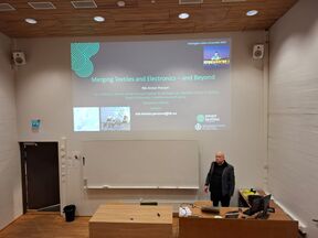 Lecturer in a modern auditorium presenting a slide titled “Merging Textiles and Electronics – and Beyond”.