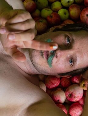 A person lies on a bed of apples, reaching out with their hand. The apples are red, yellow, and green.