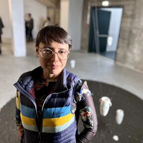 Person wearing a colourful striped vest and a patterned long sleeve shirt standing in a room with hanging white cups.
