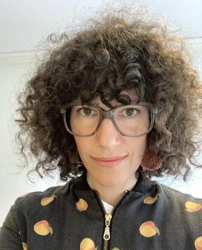 Person with curly hair wearing glasses and a dark zip-up top with peach prints.