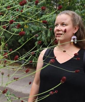 A person in a black sleeveless top is standing behind a bush with red buds. Green foliage is visible in the background.