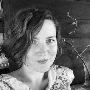 A person with wavy hair, wearing a floral patterned garment sits next to a wooden wall with a plant in a vase.