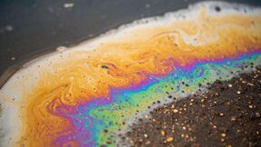 Close-up of rainbow-coloured oil slick swirling on dark, dirty water surface with floating specks