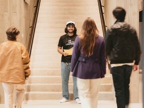 Student in black Aalto University shirt stands on indoor stairs facing three approaching people