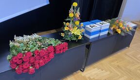 Table with red roses, colourful bouquets and blue‑white A! booklets on a wooden floor