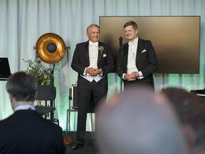 Two men in black tailcoats stand on stage by a microphone, speaking to a seated audience indoors.