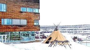 Modern wooden building beside a tan Sami tent on snowy ground, with a distant treeline