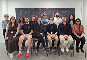 Group of people posing indoors in two rows, some seated on stools, chalkboard behind