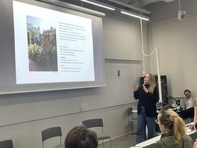 Maiju Suomi giving a lecture in a grey classroom, gesturing towards a large slide with text and a garden photo.