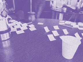 Workshop photo digitally modified with a purple duotone effect. The picture shows a table with sticky notes, a pen, a phone, a cup, thermos and mate, with people and other table in the background.