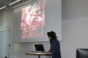 Person in dark clothes presents a colourful bedroom photo on a large white projection screen in a classroom.