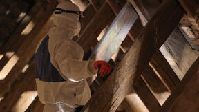 Worker in white protective gear and red gloves inspects attic roof beams.
