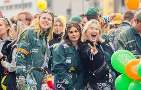 Students in green overalls at a lively outdoor event, posing for photos among orange and green balloons