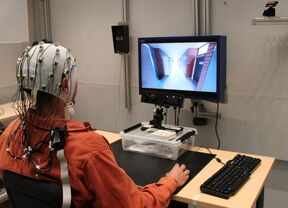 Henkilö, jolla on EEG-päähine elektrodeineen, istuu pöydän ääressä ja katsoo näytöltä virtuaalilaboratoriota. Näytön alla on silmänliikekamera.