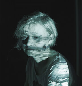 Person with light hair in a dark room, face obscured, bluish light projected on white shirt