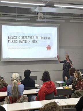 Lecture on artistic film research, presenter gesturing beside large projected slide in bright classroom.