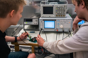 Two students test a small circuit board using oscilloscopes and grey lab instruments on a bench