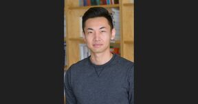 Person with blurred face in a dark grey top standing indoors in front of wooden bookshelves.