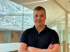 Person with blurred face in dark polo shirt, arms crossed, standing in bright modern glass office