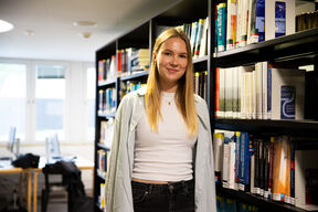 Person stands between tall library bookshelves in a bright room with computers behind