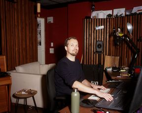 Person at mixing desk in a dim recording studio with wooden panels, computer screens and microphones