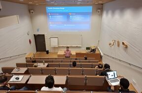 Man giving lecture on textile chemistry in tiered auditorium, students seated with laptops and notes