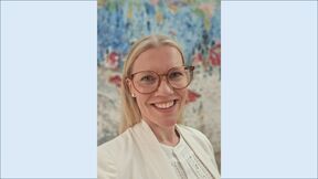 Person in cream blazer and glasses stands indoors before a colourful abstract wall painting