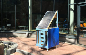 Mobile blue cart with tilted solar panel outside a glass-walled building on a brick courtyard