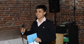 Smiling young man speaking into a microphone, holding blue papers, standing by brick wall indoors