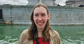 Smiling young woman in tan coat by green fountain pool with stone sculptures in the background