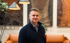 Vertics founder smiling at the camera. White man wearing a dark sweater. In the background, different pairings and lights and a sofa.