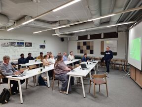 People seated at long white tables with laptops as a man presents slides in a modern classroom.