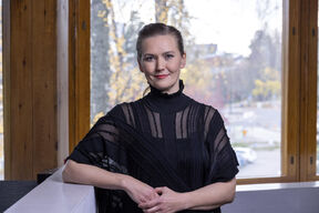 Musta-asuinen nainen nojaa valkoiseen kaiteeseen sisällä, taustalla syksyinen kaupunkimaisema ikkunoista