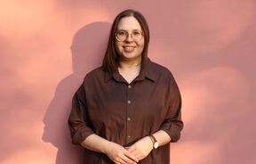 Smiling person in glasses and brown shirt stands against a pink wall in warm light