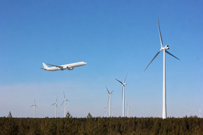 Valkoinen matkustajakone lentää metsän ja korkeiden valkoisten tuulivoimaloiden yllä sinisellä taivaalla