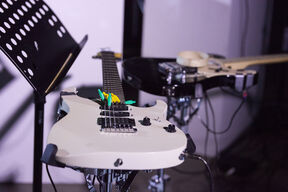 White electric guitar with colourful picks on strings, next to a black guitar and music stand on a dim stage.