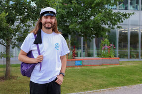 Jacek Pawlak with a student cap and purple backpack stands on green campus lawn by trees and modern building