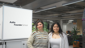 Two young women stand smiling in a modern Aalto Founder School office, with plants and desks behind them.