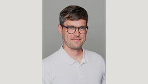 Smiling man with short brown hair and glasses in a white polo shirt, against a plain grey background