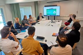 People seated around a white table in a meeting, watching a laptop presentation on a large screen.