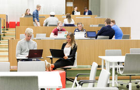 Students working on laptops in a tiered, modern classroom with light wood and grey chairs