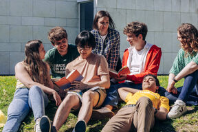 Group of students sitting on grass outside a building, reading books and chatting in the sun