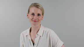 Smiling woman with light hair in a cream shirt, standing against a plain light grey background
