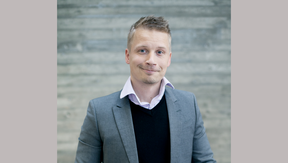 Smiling man in grey suit, black jumper and pale shirt, standing before a concrete wall