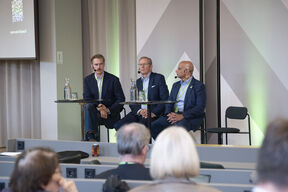 Heikki Lempinen, Jukka Salonen and Munir Mandviwalla with microphones sit at a panel table, speaking to an audience in a modern hall.