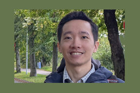 Smiling person in a jacket outdoors on a tree-lined path with green foliage in the background