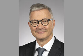 Smiling middle‑aged man in dark suit and tie, round black glasses, short grey hair, neutral grey background